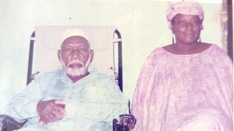 L'image montre un homme et une femme assis côte à côte. L'homme, plus âgé, est en fauteuil roulant et porte une tenue traditionnelle, comprenant un turban blanc. Il a une barbe grise et semble contemplatif. À côté de lui, la femme, qui a également des traits traditionnels, est vêtue d'une robe fluide et d'un foulard. Elle a une expression douce et chaleureuse. Le fond de l'image est sobre, créant un contraste avec leurs vêtements. L'atmosphère dégage une sensation de respect et de sérénité.