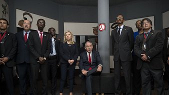 L'image montre un groupe de personnes rassemblées dans une salle. Au centre, un homme est assis sur un canapé, vêtu d'un costume, tandis que les autres se tiennent debout autour de lui. Ils portent tous des costumes et ont des expressions sérieuses, suggérant une atmosphère formelle. L'environnement semble être une exposition ou un espace culturel, avec des œuvres d'art ou des objets exposés sur les murs. L'éclairage est doux, créant une ambiance réfléchie et attentive.