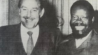 L'image montre deux hommes se tenant côte à côte, souriant largement. Le premier homme, à gauche, porte un costume sombre avec une cravate. Il a des cheveux grisonnants, une moustache bien dessinée et une expression amicale. Le deuxième homme, à droite, est habillé d'une veste de style militaire et a une barbe bien fournie. Il a les cheveux frisés et sourit également, ce qui donne une atmosphère chaleureuse à la photo. Les deux tiennent des verres à la main, suggérant une occasion conviviale, peut-être une célébration ou un événement. L'arrière-plan est flou, mettant l'accent sur les deux hommes.