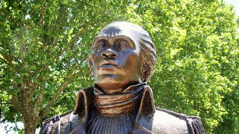 Voici la description de l'image : L'image montre une statue en bronze représentant un personnage en buste, probablement une figure historique. Le buste a un regard tourné vers le haut, suggérant une attitude déterminée ou réfléchie. Le personnage porte une veste à col haut, avec un plissé distinct sur le devant. Les détails du visage, bien que stylisés, montrent une expression forte, avec des traits bien définis. La statue est placée à l'extérieur, entourée d'arbres verdoyants, créant un cadre naturel agréable. La lumière du soleil illumine la statue, accentuant ses contours et sa texture.