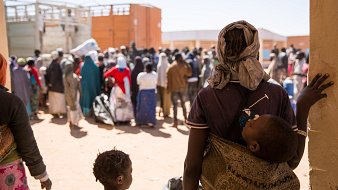 L'image montre une scène poignante dans un contexte de rassemblement de personnes, probablement dans un camp ou un centre d'accueil. Au premier plan, une personne, probablement une mère, porte un enfant dans un sling. Elle regarde vers un groupe de personnes, qui attendent patiemment, formant une masse compact de silhouettes. La majorité des personnes dans le fond sont habillées de vêtements aux couleurs variées, certaines portant des foulards ou des vêtements traditionnels. Le sol est en terre battue, et des containers peuvent être aperçus à l'arrière-plan, suggérant un environnement temporaire. L'atmosphère semble à la fois calme et chargée d'une certaine tension, reflétant une réalité complexe.