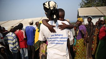 L'image montre un rassemblement de personnes dans un environnement de camp. Au premier plan, une femme, vue de dos, porte un t-shirt blanc portant une inscription en bleu qui dit : « Les Réfugiés : des personnes ordinaires traversant des moments extraordinaires. » Dans ses bras, elle tient un enfant, qui semble se blottir contre elle. En arrière-plan, on peut apercevoir un groupe de personnes, principalement des adultes, ayant des expressions variées. L'ensemble de la scène évoque des thèmes de vulnérabilité et de solidarité. Les vêtements des personnes présentes sont colorés, et l'atmosphère semble à la fois tendue et solidaire. Le décor suggère un campement temporaire, probablement en réponse à une crise.