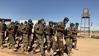 Dans cette image, un groupe de soldats est en train de marcher en formation sur un chemin poussiéreux. Ils portent des uniformes militaires verts camouflés et des casquettes vertes. La majorité d'entre eux portent également des masques bleus, ce qui suggère une mesure de protection sanitaire. En arrière-plan, on peut distinguer des conteneurs et une tour d'eau. Le ciel est dégagé, avec une couleur bleue intense, créant un contraste avec la terre orange et les silhouettes des soldats. L'atmosphère évoque un moment de discipline et d'organisation dans un environnement militaire.