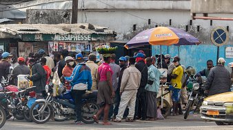L'image montre une scène animée dans une rue d'une ville. On peut voir un groupe de personnes rassemblées autour d'un kiosque à journaux, qui semble être un lieu de rencontre. Certaines personnes portent des masques, ce qui indique une attention à la santé publique. À l'avant, une femme porte un grand panier de bananes sur sa tête, ce qui met en valeur une pratique courante dans de nombreuses cultures. Des motos et une voiture jaune sont garées à proximité, ajoutant à l'ambiance urbaine. En arrière-plan, on peut distinguer des bâtiments simples, avec des couleurs plutôt ternes, ce qui donne un aspect de vie quotidienne dans ce quartier.