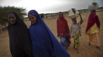 L'image représente un groupe de cinq femmes marchant sur un chemin de terre. Elles portent des vêtements traditionnels colorés, avec des motifs variés. Certaines femmes ont un voile couvrant leur tête, et une jeune fille à l'arrière transporte un objet sur sa tête, probablement un panier. Le paysage environnant est sec et désertique, avec quelques arbustes épars en arrière-plan, et le ciel est couvert, suggérant une ambiance calme et sereine. L'ensemble transmet une impression de solidarité et de culture, avec un accent sur les traditions vestimentaires.