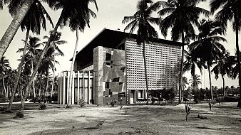 L'image montre un bâtiment moderne, avec une architecture distincte et géométrique. Le mur de la façade est ornée de motifs en relief, créant un jeu d'ombres et de lumière. Des colonnes blanches se dressent devant l'entrée principale, ajoutant une touche de classicisme à la structure. Autour du bâtiment, des palmiers élancés se balancent doucement, donnant une ambiance tropicale. Le sol est nu, avec une végétation assez faible, ce qui permet de mettre en valeur l'architecture. La scène semble baignée dans une lumière naturelle, évoquant une atmosphère calme et sereine.