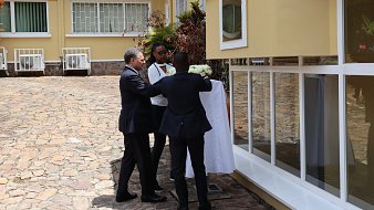 Dans cette image, nous voyons un événement en extérieur, probablement une cérémonie de dévoilement. Trois hommes sont présents. L'un d'eux, portant un costume sombre, est en train de tirer une couverture blanche qui cache un objet ou une plaque. À ses côtés, un homme en tenue décontractée tient un bouquet de fleurs. En arrière-plan, on peut apercevoir un bâtiment avec de grandes fenêtres et une pelouse soignée, créant une atmosphère accueillante. Le sol est en pierre, ajoutant un aspect naturel à l'environnement. L'ensemble de la scène évoque un moment de célébration ou d'inauguration.