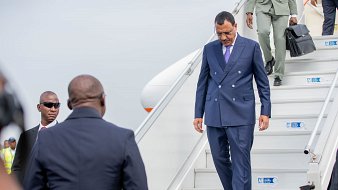 L'image montre un homme qui descend des marches d'un avion. Il porte un costume bleu foncé et des lunettes de soleil. Son expression semble concentrée tandis qu'il s'avance avec assurance. À côté de lui, un autre homme en costume noir observe attentivement. En arrière-plan, on aperçoit des membres du personnel de l'aéroport, ainsi que l'avion stationné. L'atmosphère peut sembler formelle et dynamique, suggérant un moment important, vraisemblablement une arrivée ou une sortie officielle.