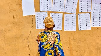L'image montre un homme de dos, vêtu d'une robe colorée avec des motifs traditionnels, principalement en bleu et jaune. Il se tient devant un mur de couleur ocre, où sont affichés plusieurs documents. Ces documents ressemblent à des bulletins de vote ou des listes, alignés verticalement, avec des photographies et des noms inscrits. Le sol est légèrement fissuré, ajoutant une texture au fond de l'image. L'homme porte un turban beige, et son attention semble focalisée sur les informations affichées sur le mur.