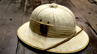 L'image montre un chapeau de style explorer, souvent associé aux expéditions dans des régions chaudes ou tropicales. Le chapeau est de couleur claire, presque beige, avec une large bordure qui protège du soleil. Sa partie supérieure est légèrement surélevée, indiquant une forme arrondie. À l'avant, il y a une bande de tissu plissé avec un morceau de bois ou de cuir, donnant un aspect rustique. Une fine tige est posée sur le chapeau, ajoutant une touche naturelle. La surface du chapeau semble légèrement usée, témoignant d'un usage antérieur. Le fond sur lequel il repose est en bois, ce qui renforce l'atmosphère d'aventure.