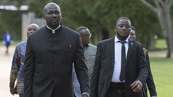 L'image montre un groupe d'hommes marchant ensemble sur un chemin dans un environnement verdoyant. Au premier plan, un homme grand et imposant porte un costume noir avec un col Mao, tandis qu'un autre homme à côté de lui est en costume noir et cravate. Ils ont l'air sérieux et concentrés, comme s'ils se rendaient à un événement important. En arrière-plan, d'autres hommes sont visibles, avec des tenues variées, ce qui suggère une atmosphère de réunion ou de conférence. Le paysage est paisible, avec des arbres et une lumière douce, ajoutant à l'ambiance formelle de la scène.