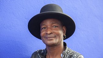 Cette image montre un homme souriant, portant un grand chapeau noir qui a une forme de haut de forme. Il est habillé d'une veste en cuir noire ornée de clous, ce qui lui donne un style à la fois décontracté et audacieux. L'arrière-plan est d'un bleu vif, créant un contraste frappant avec ses traits. Son expression est amicale et accueillante, évoquant une personnalité chaleureuse et charismatique.
