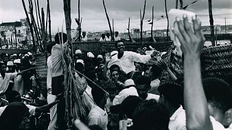 L'image montre une scène animée et dynamique, probablement lors d'un rassemblement ou d'une célébration. De nombreuses personnes se regroupent, leurs visages expriment la joie et l'excitation. On peut voir des hommes et des femmes, certains portant des vêtements traditionnels. Au centre, un homme semble chercher à atteindre un groupe, suscitant des réactions enthousiastes de la foule. Les bras de plusieurs personnes s'élèvent, clairement engagés dans l'événement. L'arrière-plan apparaît flou, avec des éléments de construction ou des arbres qui ajoutent à l'atmosphère vivante de la scène. L'ambiance générale évoque une forte connexion et une communauté vibrante.
