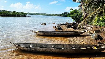 Imaginez une scène paisible au bord de l'eau. Devant vous se trouve un rivage doux avec du sable doré, illuminé par le soleil. À votre gauche, deux pirogues en bois, usées par le temps, reposent tranquillement sur le rivage, légèrement inclinées vers l'eau calme. La surface de l'eau est lisse, reflétant le ciel bleu parsemé de quelques nuages blancs. À droite, vous pouvez percevoir des rochers de tailles variées, surmontés ici et là de végétation luxuriante. Des palmiers se dressent en arrière-plan, ajoutant une note tropicale à l'ambiance. L'air est frais, avec une légère brise qui apporte une odeur de terre humide et de végétation. L'ensemble dégage une atmosphère de sérénité et de beauté naturelle, propice à la contemplation.
