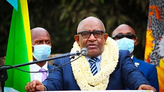 Sur cette image, un homme est debout devant un microphone, en train de s'exprimer. Il porte un costume sombre avec une chemise claire et une cravate rayée. Autour de son cou, une couronne de fleurs blanches et jaunes symbolise un accueil ou un hommage. En arrière-plan, plusieurs personnes l'écoutent attentivement, certaines portant des masques de protection. À gauche, on peut voir des drapeaux aux couleurs vives qui ajoutent une touche festive à la scène. L'ambiance semble sérieuse et solennelle.