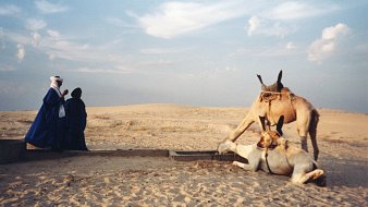 L'image montre un paysage désertique, caractérisé par des dunes de sable qui s'étendent à perte de vue sous un ciel légèrement nuageux. Au premier plan, deux personnes, habillées de vêtements traditionnels, se tiennent près d'un point d'eau. L'une d'elles, en bleu, semble prier ou méditer. À proximité, un chameau repose tranquillement sur le sol, tandis qu'un autre chameau se penche pour boire. L'ambiance de la scène est paisible, avec une lumière douce qui souligne les formes des dunes et des silhouettes. Les sons du désert, comme le vent léger, pourraient renforcer cette atmosphère sereine.