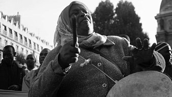 L'image représente une femme engagée dans un moment de mobilisation. Elle porte un manteau gris et un foulard autour de la tête. Ses bras sont levés, tenant des baguettes, et elle semble jouer d'un instrument de percussion, probablement un tambour. Son visage exprime une concentration et une passion profonde, alors qu'elle fait vibrer la musique au sein d'une foule. En arrière-plan, on peut distinguer d'autres personnes, suggérant une manifestation ou un rassemblement collectif. L'atmosphère est chargée d'énergie et de détermination. La photo est en noir et blanc, ce qui ajoute une dimension intemporelle à cette scène de citoyenneté active.