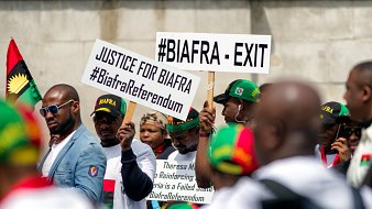 L'image montre un groupe de personnes participant à une manifestation. Elles portent des t-shirts blancs et des casquettes aux couleurs du mouvement Biafra. Des pancartes sont brandies, portant des inscriptions telles que "#BIAFRA - EXIT" et "JUSTICE FOR BIAFRA", ainsi que des hashtags liés au référendum biafrais. L'atmosphère semble dynamique et engagée, reflétant une revendication pour la justice et l'autodétermination.