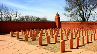 L'image montre un espace extérieur bien entretenu, avec plusieurs bornes ou stèles de forme allongée disposées en rangées sur un sol en gravier. Le sol est également pavé en terrasse, et il y a un mur de couleur beige orangé qui entoure la zone. En arrière-plan, des arbres dénudés se dressent contre un ciel bleu clair avec quelques nuages. L'ensemble dégage une atmosphère de sérénité et de réflexion.