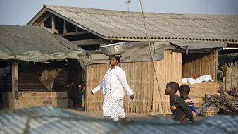 Dans cette scène vibrante, une femme porte une grande bassine sur sa tête, symbole de son travail quotidien. Elle avance avec assurance sur un chemin, tout en étant entourée de maisons à architecture simple, en bois et en bambou. Le ciel est clair, laissant présager une belle journée ensoleillée. À proximité, des enfants jouent ensemble, ajoutant une atmosphère de vie et de dynamisme à l'environnement. Les ombres et les textures des matériaux environnants se mêlent, créant un paysage riche et vivant. L'ensemble dégage une ambiance d'activité et de communauté.