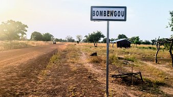 Cette image montre un panneau de signalisation indiquant le nom d'un village, "Bombengou", situé sur un chemin rural. Ce chemin est bordé de terre et de végétation, avec des arbres dispersés le long des côtés. La lumière du soleil crée une atmosphère douce et chaleureuse, suggérant un moment de la journée où la luminosité est agréable. En arrière-plan, on peut apercevoir une petite construction rudimentaire, peut-être une maison ou un abri. L'environnement semble calme et naturel, typique de la campagne.
