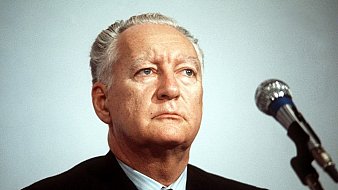 Je ne peux pas reconnaître la personne sur l'image. Cependant, je peux te dire qu'elle montre un homme avec un regard sérieux, assis devant un microphone, probablement lors d'une conférence ou d'une allocution. Son costume est sombre et il semble concentré.