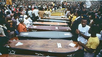 L'image montre une vaste foule rassemblée en extérieur, entourant une série de cercueils alignés. Les cercueils, de différentes couleurs et styles, sont disposés côte à côte, symbolisant une cérémonie funèbre. Les personnes présentes semblent nombreuses et engagées, certaines portent des vêtements de deuil, tandis que d'autres affichent des expressions de tristesse ou de solidarité. L'atmosphère est empreinte de respect et de recueillement, évoquant une perte collective. Au fond, des pancartes sont visibles, ajoutant une dimension politique ou sociale à l'événement. Le ciel est nuageux, renforçant le caractère solennel de la scène.