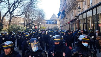 L'image montre une scène urbaine animée. En premier plan, un groupe de policiers en uniforme, équipés de casques et de protections, se tient rassemblé. Leur posture est attentive, suggérant une opération en cours ou une manifestation. À l'arrière-plan, des arbres dénudés et des bâtiments historiques sont visibles, indiquant que la scène se déroule probablement dans une zone urbaine. C'est une journée ensoleillée, avec un ciel clair, apportant une lumière vive sur la scène. L'atmosphère semble tendue, avec des gens qui observent la situation depuis les côtés. On peut percevoir une impression d'encadrement, de sécurité, mais aussi de tension sociale.