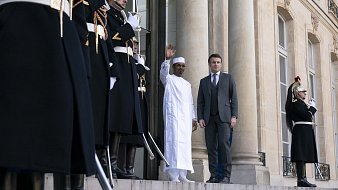 L'image montre une scène officielle à l'extérieur d'un bâtiment majestueux. Deux hommes se tiennent sur les marches devant l'entrée principale. À gauche, un homme porte une tenue traditionnelle blanche, levant la main en salutation. À sa droite, un homme en costume sombre regarde légèrement vers lui. En arrière-plan, plusieurs gardes d'honneur, habillés de costumes noirs avec des ornements dorés et des plumes rouges sur leurs casques, se tiennent en position, ajoutant à l'atmosphère solennelle de la scène. Les colonnes imposantes du bâtiment et les détails architecturaux mettent en valeur le caractère formel de l'événement.