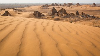 L'image décrit un paysage désertique, dominé par de vastes dunes de sable aux formes ondulantes. En arrière-plan, on aperçoit des pyramides anciennes dont les sommets sont pointus et les surfaces sont qui semblent rugueuses. Le terrain autour des pyramides est également parsemé de petites éboulis et de dunes, créant un paysage à la fois majestueux et mystérieux. La lumière du soleil éclaire la scène, mettant en valeur les nuances dorées du sable et l'architecture historique des pyramides. L'atmosphère générale évoque la chaleur et l'immensité du désert, ainsi qu'un sentiment d'émerveillement face aux vestiges du passé.