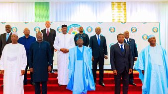 L'image montre un groupe de plusieurs hommes posant ensemble sur une scène. Ils sont vêtus de costumes formels et de tenues traditionnelles. La scène est décorée avec un fond de couleurs vives, comprenant un motif de logo sur la toile blanche. Les hommes se tiennent sur une estrade de couleur rouge, formant une sorte de demi-cercle. Certains portent des vêtements à motif et d'autres des costumes classiques, reflétant une diversité culturelle. L'atmosphère semble officielle, suggérant un événement important ou une réunion.