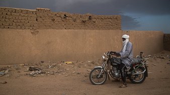 L'image montre un homme immobile à côté de sa moto dans un paysage désertique. Il porte un long vêtement traditionnel et un turban qui couvre sa tête, laissant seulement ses yeux visibles. Le sol est constitué de sable et de débris, tandis qu'en arrière-plan, on distingue un mur de terre crue, simple et robuste, typique des constructions dans cette région. Le ciel est nuageux, ajoutant une atmosphère dramatique à la scène. L'homme semble contemplatif, connectant ainsi l'élément humain avec l'environnement désertique qui l'entoure.