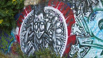 L'image représente une peinture murale vibrante sur un mur en béton. Au centre, on peut voir une figure stylisée, habillée de manière étrange, qui semble exprimer une sorte de défi ou de résistance avec des gestes des mains. La figure est entourée d'un motif circulaire qui est accentué par des couleurs vives comme le rouge et le noir, créant un fort contraste visuel. D'autres graffitis colorés sont visibles autour, ajoutant une diversité de formes et de couleurs à l'ensemble. À la base, des feuilles vertes, provenant de la végétation environnante, sortent du sol, créant une atmosphère vivante et urbaine.