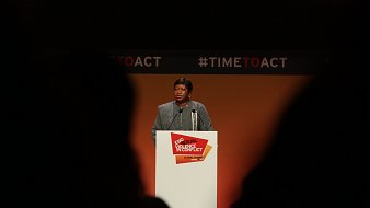 L'image montre une femme debout au micro, s'adressant à un public. Elle porte un vêtement formel et a une expression sérieuse. Le fond est principalement orange, avec des éléments graphiques en noir et rouge. Un panneau blanc devant elle affiche le texte "End Sexual Violence in Conflict" et "#TIMETOACT". La scène semble être un événement ou une conférence sur un sujet important, probablement lié à la lutte contre la violence sexuelle en contexte de conflit. Les silhouettes des personnes dans le public sont visibles en bas de l'image, ajoutant à l'ambiance de l'événement.