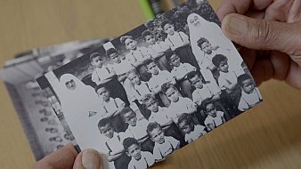 L'image montre une photographie en noir et blanc d'un groupe d'enfants, probablement d'une école ou d'une classe. Les enfants, qui portent des uniformes clairs, sont rassemblés en plusieurs rangées, affichant des expressions diverses. Au centre, une personne vêtue d'une tenue traditionnelle, probablement une enseignante ou une surveillante, se tient parmi eux. L'arrière-plan est flou, mais on peut deviner une ambiance scolaire. Les mains de quelqu'un tiennent la photo avec précaution, indiquant son importance personnelle ou sentimentale.