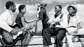 L'image représente un groupe de quatre personnes en noir et blanc, assises autour d'une barrière en bois, sous un ciel partiellement nuageux. Deux hommes et deux femmes sont présents. L'un des hommes joue de la guitare, tandis que l'autre est assis sur un tronc, tenant un instrument à cordes, probablement un banjo ou un ukulélé. Les deux femmes semblent interagir avec l'un des hommes et un cheval qui est également présent, la tête tournée vers eux. Elles affichent des expressions joyeuses, créant une atmosphère conviviale et détendue. Les vêtements des personnages varient : l'un porte un chapeau de cowboy et les autres portent des vêtements simples, typiques de l'époque. L'image évoque un moment de camaraderie et de musique, suggérant un lien fort entre les personnes et la nature environnante.