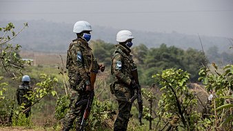 L'image montre deux militaires en uniforme, portant des casques blancs et des masques. Ils se tiennent debout sur un terrain légèrement vallonné, entourés de verdure, avec des plantes et des arbres en arrière-plan. Les soldats tiennent des armes et semblent vigilants, observant leur environnement. L'atmosphère est sérieuse, évoquant un contexte de mission ou de sécurité. Le ciel est clair, et l'ambiance générale peut donner une impression de calme malgré la présence militaire.