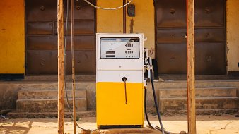 Imaginez une petite station-service située à l'extérieur d'un bâtiment. Au centre de l'image se trouve une pompe à essence de couleur blanche et jaune, installée sous un auvent en tôle ondulée qui offre un peu d'ombre. La pompe est équipée d'un écran et d'un bouton pour le paiement, ainsi qu'un flexible noir reliant la pompe au réservoir d'un véhicule. À l'arrière-plan, on aperçoit un mur de couleur jaune avec deux portes en métal marron, fermées, et l'ensemble est posé sur un sol terreux, suggérant une ambiance rustique et simple. Le lieu semble tranquille, sans voitures visibles autour de la pompe.