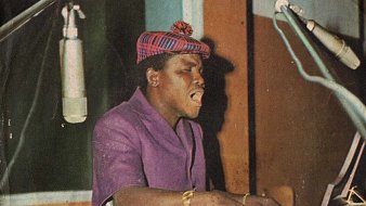 Sur cette image, nous voyons un homme en train de jouer du piano. Il porte une chemise violette et un chapeau à motif écossais. Son expression est concentrée et passionnée alors qu'il chante. Deux microphones sont placés à proximité, capturant sa voix. L'arrière-plan présente des panneaux en bois colorés, créant une ambiance chaleureuse et artistique. L'atmosphère dégage une sensation de musique vivante et d'émotion.