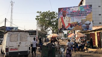 L'image représente une scène de rue animée dans une ville. Au premier plan, on voit une camionnette blanche à l'arrière, avec des détails usés et des portes ouvertes. À gauche, un bus de transport en commun est stationné. Plusieurs personnes se déplacent sur la chaussée, certaines vendant des marchandises sous des parasols colorés. Il y a également des bacs de récupération et divers déchets sur le sol, indiquant une ambiance de marché. En arrière-plan, un grand panneau publicitaire présente des visages de personnes et un message en français, affichant un slogan qui semble inciter à l'action ou au changement. Les arbres et les fils électriques ajoutent à l'atmosphère urbaine du lieu.