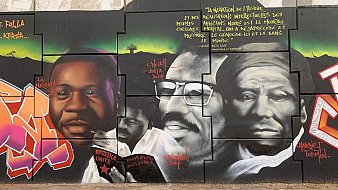 Cette image montre une fresque murale vibrante et colorée qui rend hommage à des figures importantes de l'histoire africaine. Au centre, il y a des portraits de trois personnes, chacun portant une expression sérieuse et réfléchie. Les visages sont peints en noir et blanc, mettant en valeur les détails de leurs traits. À gauche, un homme sourit légèrement, symbolisant l'espoir et la détermination. Au centre, un homme barbu porte des lunettes et affiche un regard intense, représentant la sagesse. À droite, un homme au visage marqué par le temps semble contemplatif, illustrant la profondeur de la pensée. En arrière-plan, des motifs géométriques et des couleurs riches ajoutent une dynamique à l'ensemble. Des inscriptions en lettres blanches ponctuent la fresque, partageant des pensées inspirantes sur l'identité, la culture et l'impact de ces figures dans le monde. Cette œuvre évoque une ambiance de fierté et d'émancipation culturelle.