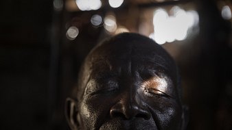 L'image montre un homme avec les yeux fermés, laissant transparaître une profonde sérénité. Son visage, marqué par le temps, présente des rides et une peau sombre. Une lumière douce filtre à travers un espace, touchant délicatement son visage et créant des reflets subtilement brillants en arrière-plan. L'ambiance de l'image est calme, évoquant un moment de méditation ou de paix intérieure. Les contours flous de l'environnement suggèrent un espace simple, sans détails distracteurs.