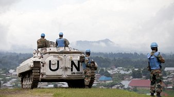 L'image présente un paysage où des soldats en uniforme de l'ONU se trouvent autour d'un véhicule blindé. Le véhicule, marqué par le symbole "UN" en lettres blanches, est stationné sur une colline qui surplombe une petite ville. Les soldats portent des casques bleu clair typiques des missions de maintien de la paix. À l'arrière-plan, on aperçoit des maisons aux toits colorés et une végétation dense, tandis que le ciel est partiellement nuageux, créant une atmosphère sereine malgré le contexte militaire. Les soldats semblent vigilants et concentrés, surveillant les environs.