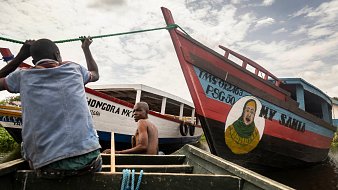 L'image montre deux bateaux colorés amarrés au bord d'un cours d'eau calme. À l'avant, un homme, en train de ramer, est visible, concentré sur son effort. Un autre homme est assis à l'arrière, regardant vers l'objectif. Les bateaux sont peints de manière vive, avec des inscriptions comme "MY SAID" et "MONGORA MK". L'un des bateaux présente également une grande peinture d'un visage souriant. Le ciel est partiellement nuageux, ajoutant une dimension lumineuse à la scène. L'atmosphère est paisible, évoquant une ambiance de vie flottante.