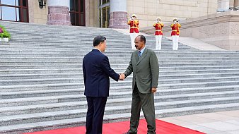 L'image montre deux hommes se serrant la main sur un tapis rouge, symbolisant un accueil officiel. Ils sont debout devant un bâtiment imposant, avec des marches qui mènent à l'entrée. En arrière-plan, on aperçoit des gardes en uniformes, debout et formant une ligne. Le premier homme porte un costume sombre, tandis que le second est habillé d'un costume vert. L'atmosphère dégage un sentiment de cérémonie et de respect mutuel.
