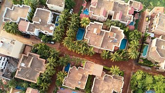 L'image montre une vue aérienne d'un complexe résidentiel. On y voit plusieurs maisons aux toits plats, disposées autour de petites piscines bleues. Parmi ces maisons, il y a de nombreuses palmiers qui ajoutent une touche de verdure. Les allées sont en terre et les maisons sont agencées de manière à créer un réseau urbain harmonieux. Les couleurs de la terre et des toits se marient pour donner une ambiance chaleureuse et accueillante au paysage.