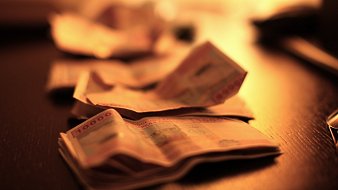 L'image montre un plan rapproché de billets de banque étalés sur une surface sombre, probablement une table en bois. Les billets sont flous, suggérant une légère profondeur de champ, ce qui attire l'attention sur la texture et les détails des billets. L'éclairage est doux et chaud, avec une lumière diffuse provenant d'une source hors du champ de vision, créant une ambiance intime et feutrée. Cette lumière fait ressortir les motifs et les couleurs des billets, ajoutant une dimension visuelle à l'ensemble.