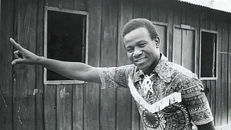 L'image montre un homme debout à côté d'une maison en bois. Il porte une chemise colorée au motif complexe, avec des nuances de marron et de beige. Son sourire est amical et engageant. L'homme pointe une main vers un mur en bois, suggérant une direction ou attirant l'attention sur quelque chose. En arrière-plan, on aperçoit des fenêtres sombres qui ajoutent une dimension de profondeur à la scène. L'atmosphère semble à la fois chaleureuse et authentique, évoquant un moment de convivialité.