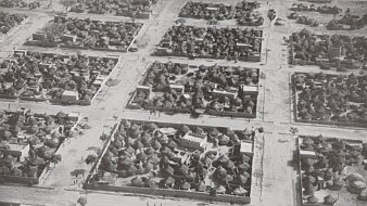 L'image montre une vue aérienne d'un village africain. On peut voir un agencement organisé des habitations, qui sont principalement des maisons traditionnelles en toit de chaume, caractéristiques de certaines régions. Les structures sont regroupées en plusieurs blocs délimités par des chemins. Des arbres sont dispersés dans l'espace, ajoutant une touche de verdure. Quelques personnes se déplacent le long des routes, suggérant une animation dans ce décor rural. Le photographique est en noir et blanc, ce qui donne une atmosphère historique à la scène.