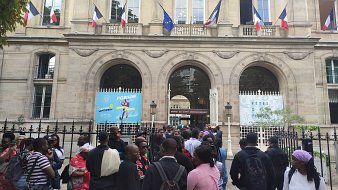 L'image montre un bâtiment imposant avec une architecture classique, visible grâce à ses grandes fenêtres et ses arches. Devant ce bâtiment, un groupe de personnes se rassemble, créant une atmosphère animée. Des drapeaux français flottent au-dessus d'eux, témoignant de l'importance de ce lieu. Les visiteurs semblent variés en termes d'âge et de style vestimentaire, ce qui reflète une diversité culturelle. À l'arrière-plan, des affiches colorées annoncent un événement, ajoutant une touche festive à la scène. L'ensemble dégage une ambiance d'attente et d'excitation.